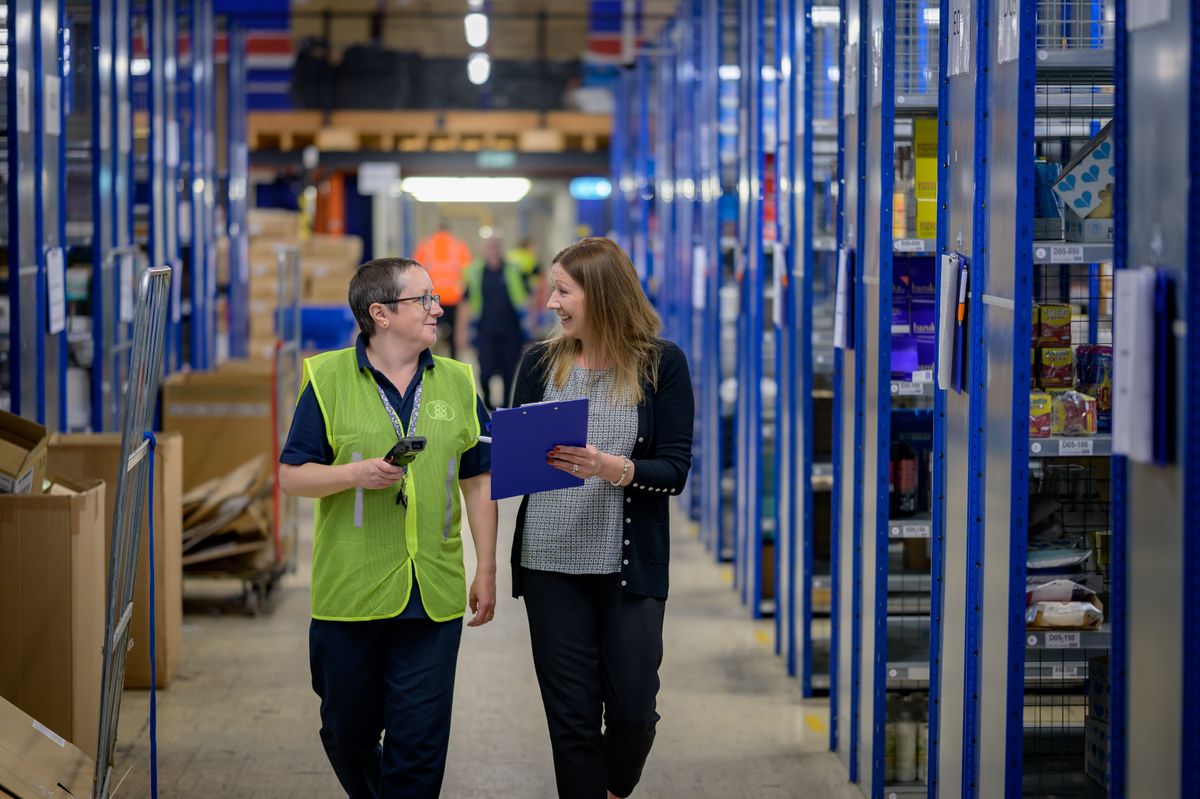 Two workers in reflective vests collaborate in a busy warehouse aisle