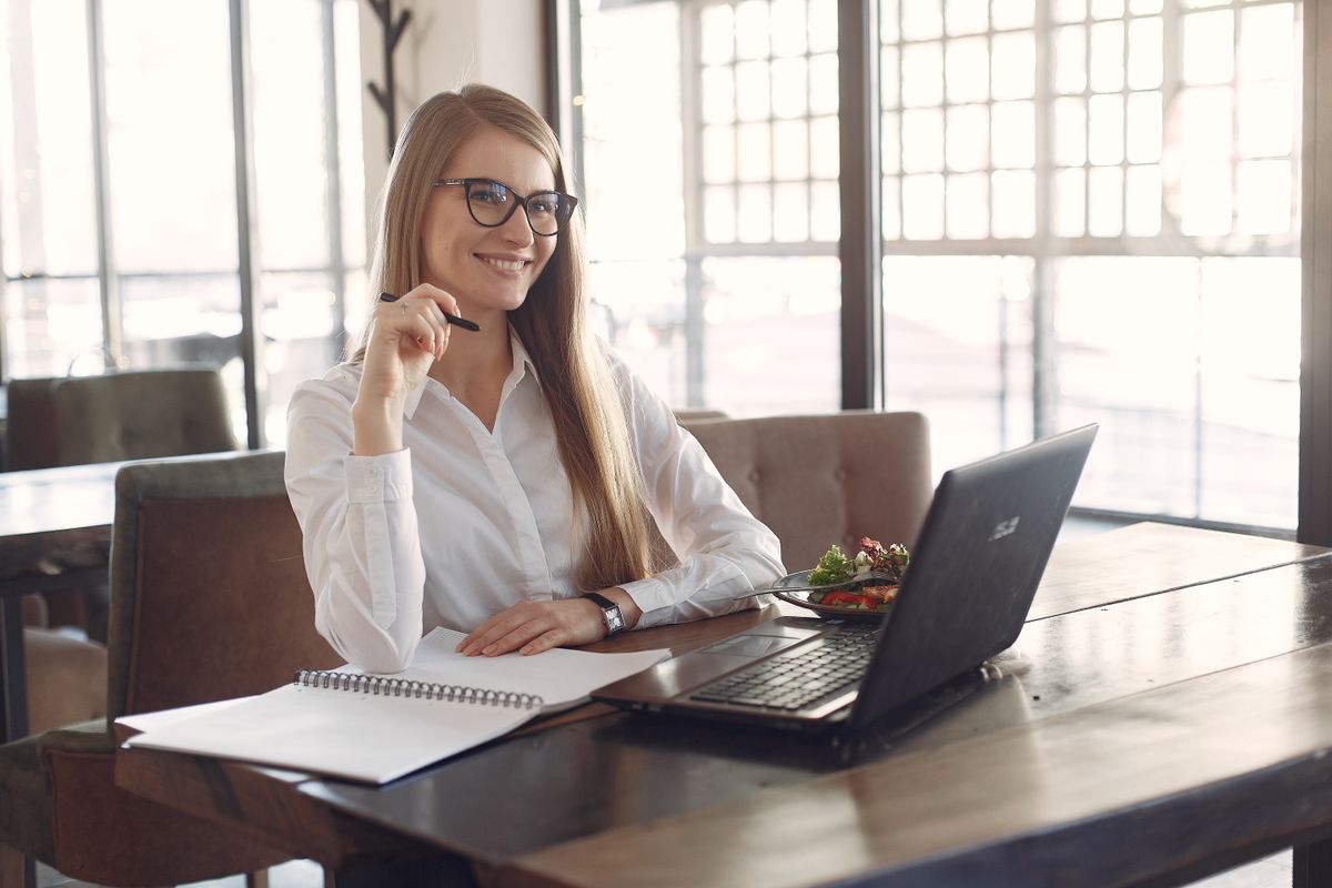 Positive smiling female entrepreneur in formal clothes and eyeglasses sitting at table with laptop and planner