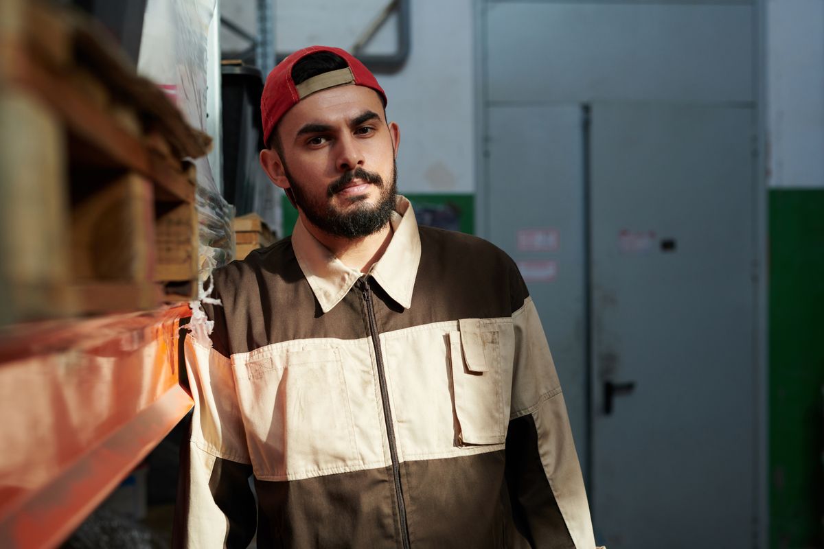 Man with beard and mustache posing in industrial warehouse setting wearing workwear